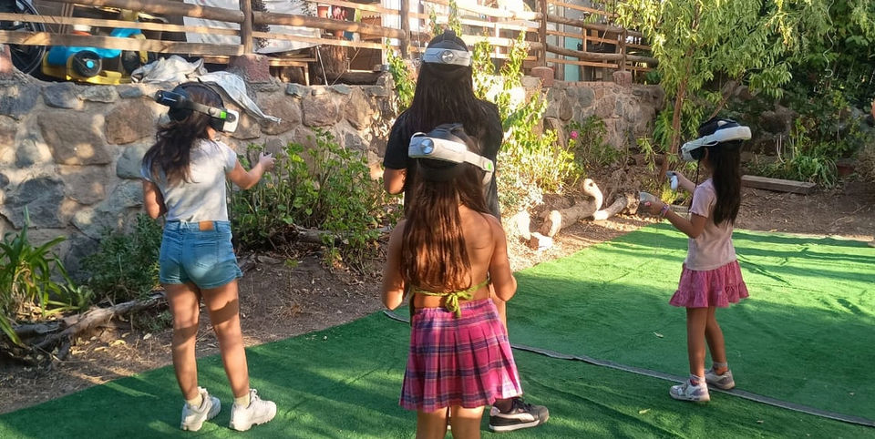 Tres niñas jugando con visores de realidad virtual inalámbricos en un jardín exterior con pasto sintético durante una fiesta de cumpleaños.