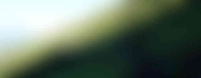 Hikers walk along a lush, green mountain trail under a bright sky. The path winds through dense foliage, with distant hills in view.