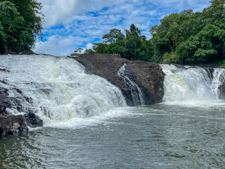 Waterfall cascading over dark rocks surrounded by lush green forest under a partly cloudy blue sky. Rippling water flows in foreground.