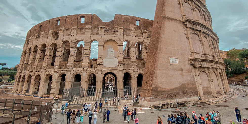 Vista panorâmica do Coliseu, imponente e parcialmente preservado, com suas paredes de pedra e arcos icônicos. No primeiro plano, turistas caminham e observam a estrutura, sob um céu nublado que dá um tom dramático à cena.