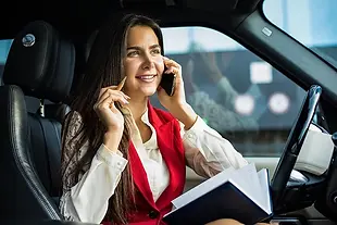 Smiling professional woman talking on phone, holding pen and notebook in car.
