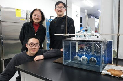 From the left: Hailong Fan (front row), Jian Ping Gong, and Hongguang Liao from the research team. Also pictured are hydrogels fixed between ceramic (left), glass (middle), and titanium (right) plates supporting 1 kg weights in normal saline for over a year. (Photo: WPI-ICReDD)