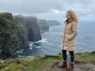 Jess standing on the Cliffs of Moher in Ireland on a cloudy winter day, overlooking the Atlantic Ocean with windblown hair and dramatic coastal cliffs behind her.