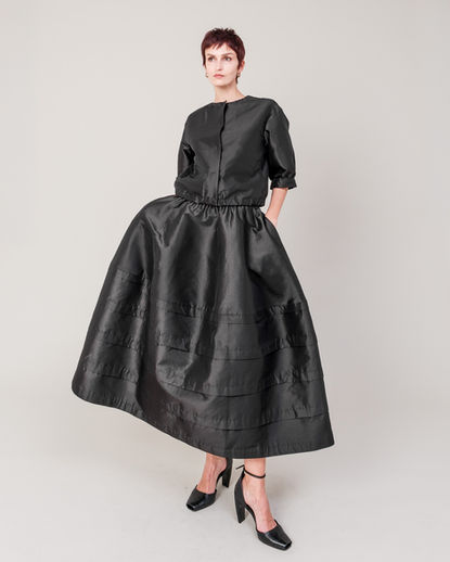 Shiny short jacket and matching shiny skirt styled as a set. The subtle reflective surface and clean, compact silhouette create a modern, structured look with gentle movement. DOMELLE 25AW look.