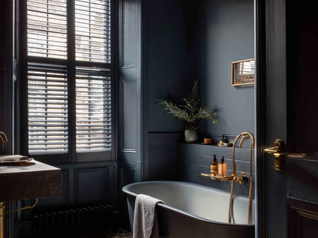 A moody, stylish East London bathroom featuring matte black plantation shutters on a tall Victorian window, deep blue panelled walls, a freestanding black bathtub with brass fixtures, patterned floor tiles, and soft natural light creating a calm, luxurious atmosphere.