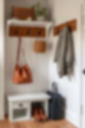 Cozy entryway with white bench, brown leather bag, shoes below, gray coat, and tote on hooks. Potted plant and basket on top shelf.