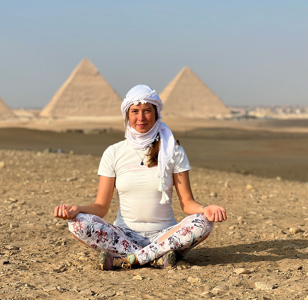 Woman meditates cross-legged in the desert before the Pyramids of Giza, wearing a white headscarf and floral leggings.