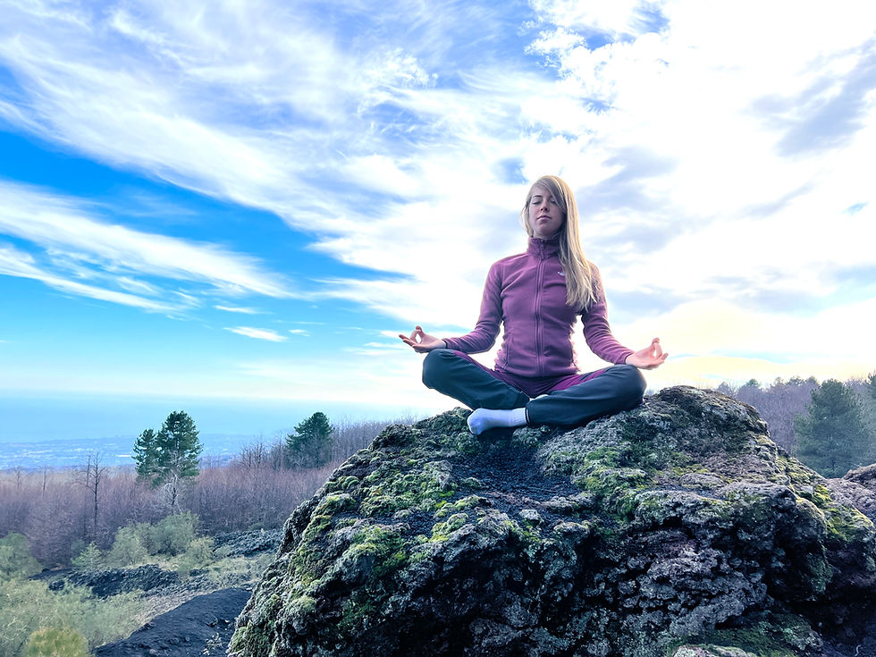 Woman in a purple jacket meditates cross-legged on a mossy rock above a forest under a bright, cloudy blue sky.