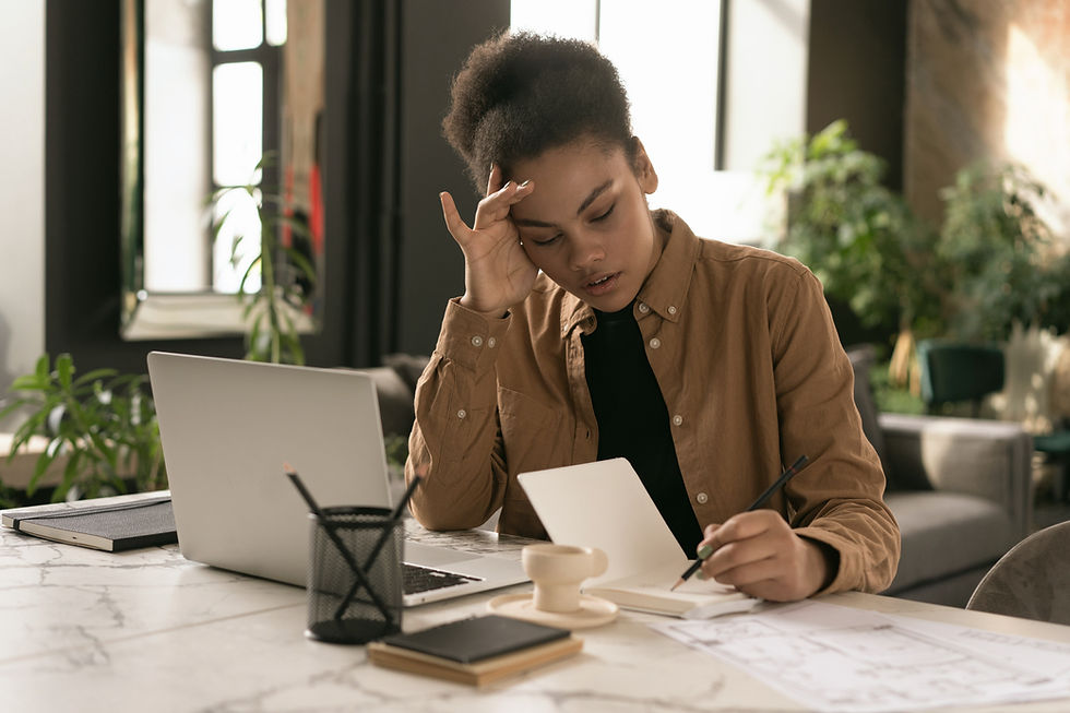 Woman pausing at a cluttered home desk, hand resting on her temple, notebook open beside her laptop in warm natural light