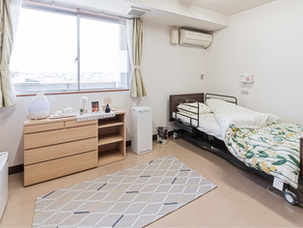 A bright, well-kept aged care room featuring a single adjustable bed with floral bedding, a wooden dresser with a humidifier and decorative items, a small TV, and a window with natural light filling the space.