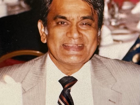 Smiling man in a beige suit and striped tie at a formal banquet, with blurred guests and table settings in the background.