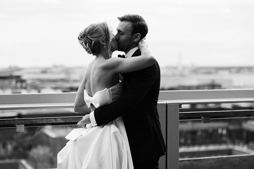 Bride and groom kissing on a rooftop. She wears a dress with a large bow, he wears a suit. City skyline in the background. Black and white.
