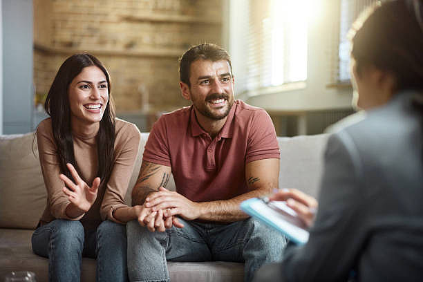 A smiling couple sits on a couch talking to a person with a clipboard in a sunlit room. The mood is warm and friendly.