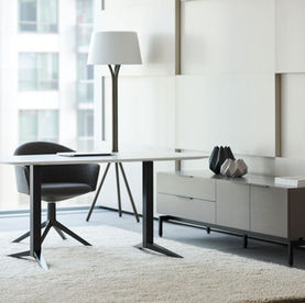 A sleek, modern office corner featuring a minimalist desk with a clean white surface and black metal legs. A dark gray upholstered swivel chair is positioned on a soft, neutral-toned rug. To the right, a low cabinet with a wood finish holds decorative geometric objects and neatly stacked magazines. Large windows offer a view of an urban building, while a contemporary floor lamp adds a touch of elegance to the space.