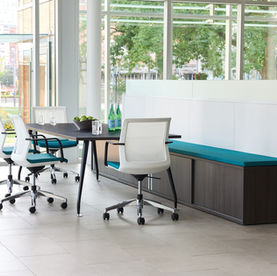 A modern office space with a black table surrounded by white office chairs, a built-in bench with teal cushions, and a large window overlooking greenery and a cityscape in the background. The table holds water bottles, glasses, and a bowl of green apples.
