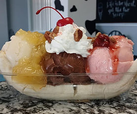 A banana split in a clear dessert dish featuring vanilla, chocolate, and strawberry ice cream topped with fruit sauces, whipped cream, chopped pecans, and a cherry.