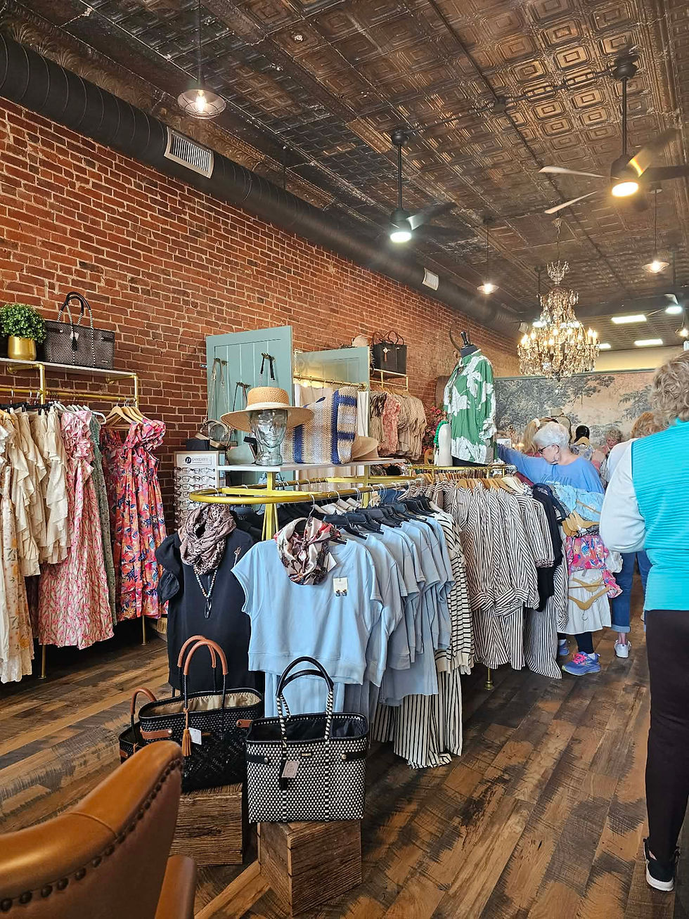 Clothing boutique with racks of colorful dresses, shirts, and scarves. Several people browse the displays under a chandelier. Exposed brick walls and decorative items like hats and handbags add to the cozy atmosphere.