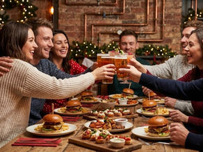 Grupo de amigos brindando con copas y cervezas sobre una mesa de madera con hamburguesas gourmet y tapas variadas, en un ambiente cálido con luces navideñas desenfocadas al fondo. Estilo vintage-industrial.