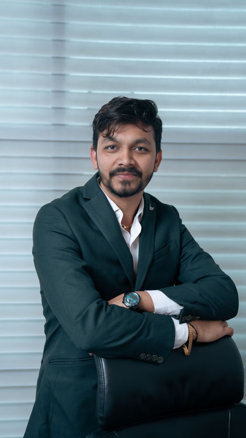 Nikhil Nimavat - Director at Heaven Investment posing confidently in a dark suit, leaning on the back of a chair in front of window blinds.