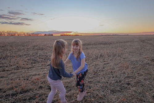 twin girls holding hands and spinning around in a farmers field with a beautiful blue, orange and pink sunset behind them.