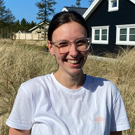 Ruth hat dunkelbraune Haare zurückgebunden und trägt eine durchsichtige Brille. Sie trägt ein weißes Tshirt und lacht. Im Hintergrund sieht man ein schwarzes Haus, einen Baum und Gräser.