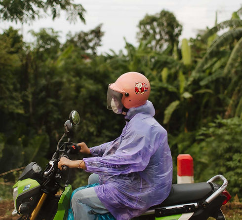Woman riding scooter beside rainforest, wearing a peach Hello Kitty helmet and a purple raincoat