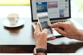 Hands holding a smartphone with a news article, tapping screen. Coffee cup on table and blurred laptop in background. Calm setting.