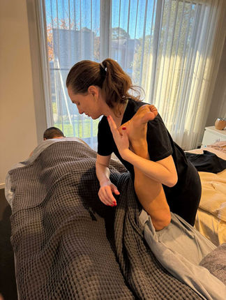 Massage therapist performing a deep tissue massage on client’s leg in a well-lit, serene room with large windows and a gray blanket.