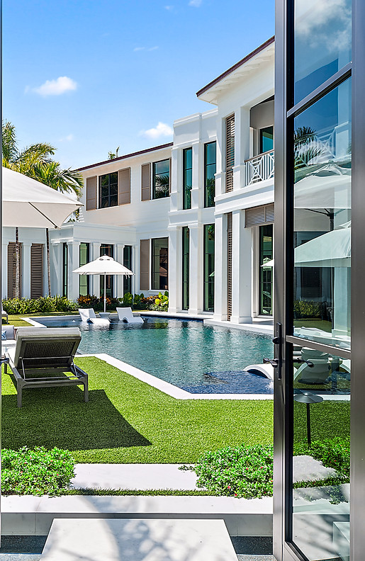 looking out to a luxurious backyard with a sparkling blue pool with lounge chairs on a sun shelf, green grass, and guest home