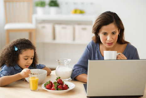 Mother at table in front of her laptop. Her child is eating breakfast next to her.