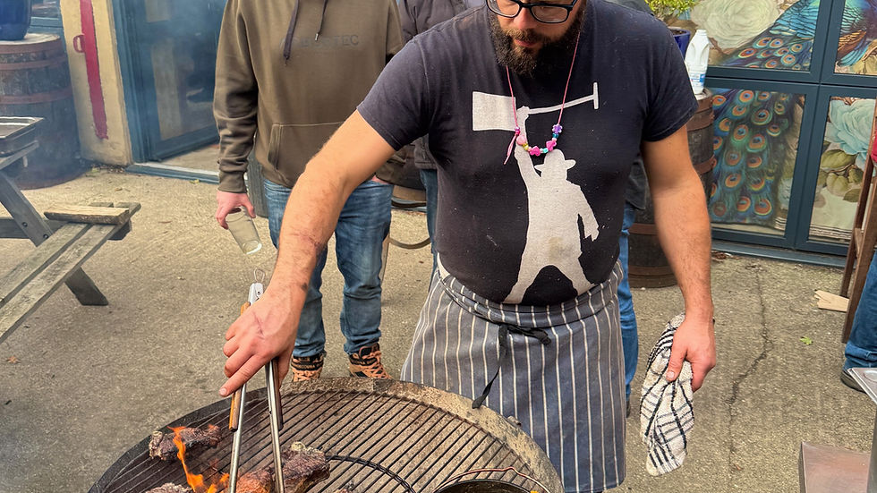 Man grilling meat and vegetables on a round barbecue. Two people stand nearby, smiling. Brick wall and glass doors in the background.