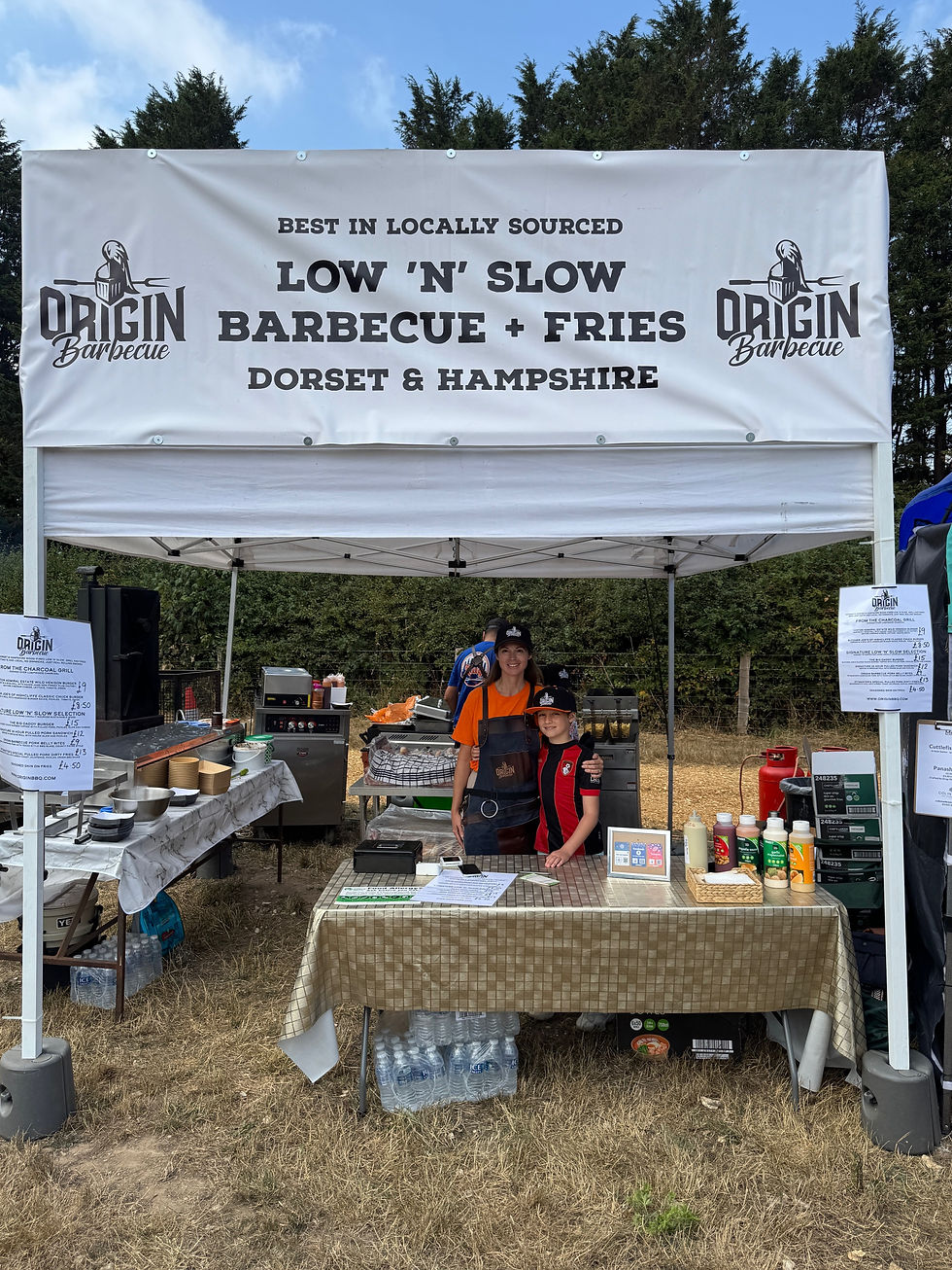 Mother and son in a BBQ stall with a sign "LOW 'N' SLOW BARBECUE + FRIES." They stand behind a table with sauces and menus. Trees in the background.