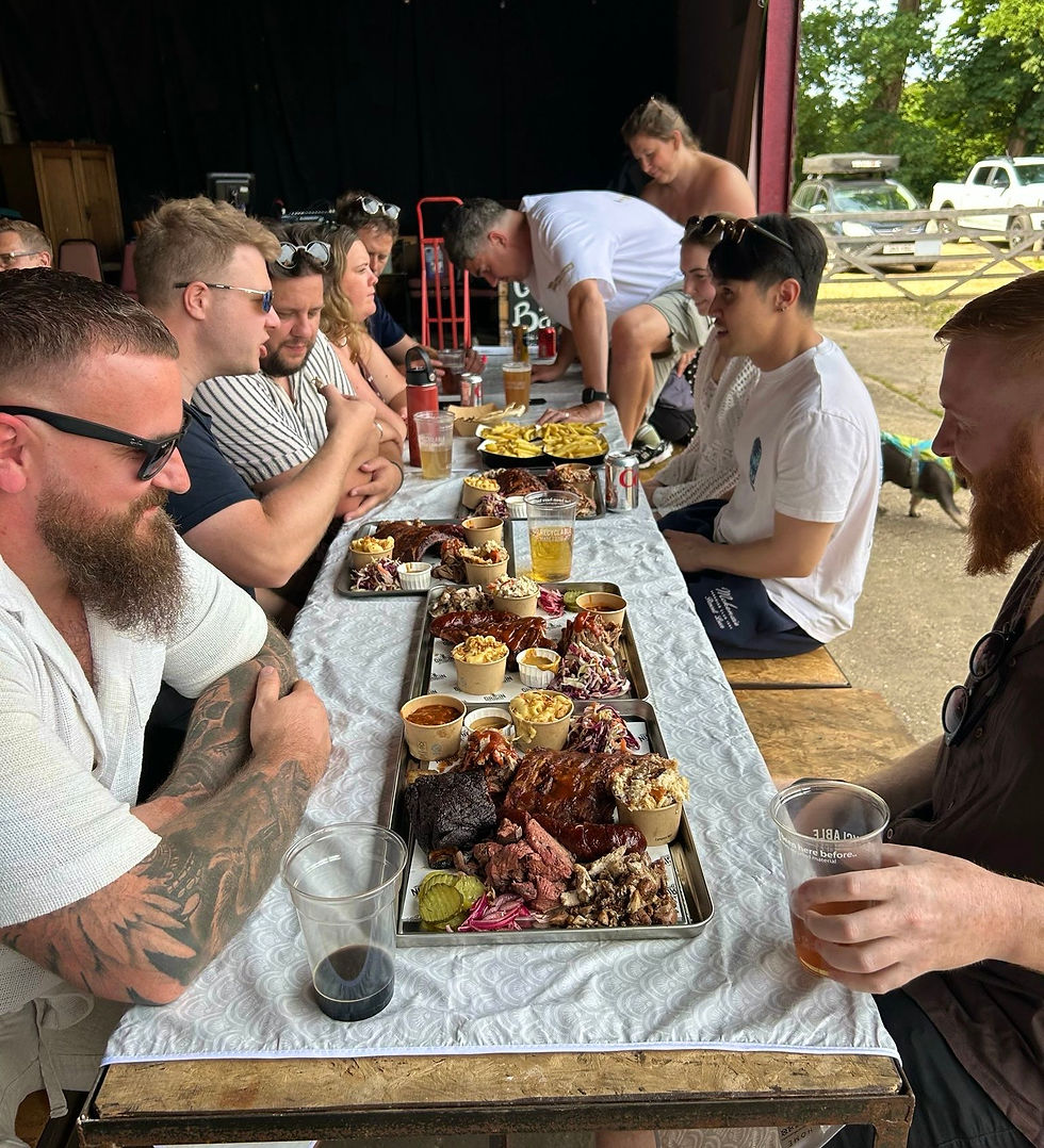 Group eating barbecue at an outdoor picnic table, with trays of meat, fries and drinks under a shaded pavilion.