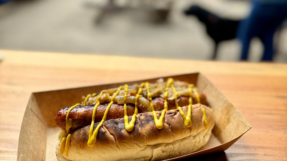 Hot dog with mustard and onions on a wooden table. Blurred background with people sitting and a black dog, creating a casual outdoor vibe.