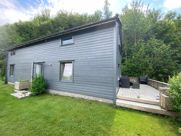 Ein graues Ferienhaus in schöner, ruhiger Umgebung mit Holzterrasse und Gartenmöbeln.