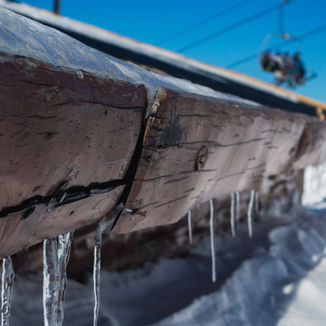 Icicles hang from a weathered wooden beam, contrasting with the bright blue sky and snow-covered landscape in the background.