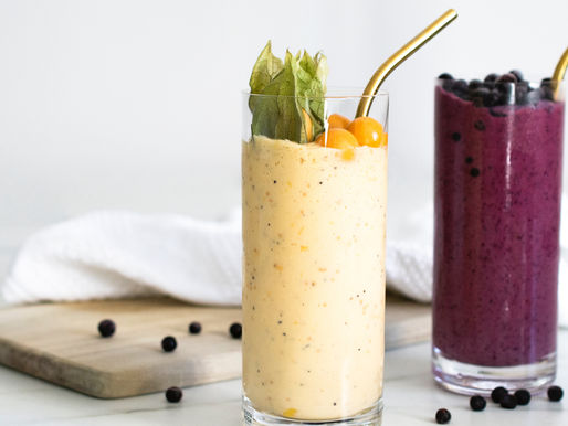 Two refreshing smoothies, one vibrant yellow garnished with physalis and the other a deep berry color with a sprinkle of blueberries on top, are served in elegant glasses with gold straws on a marble surface.