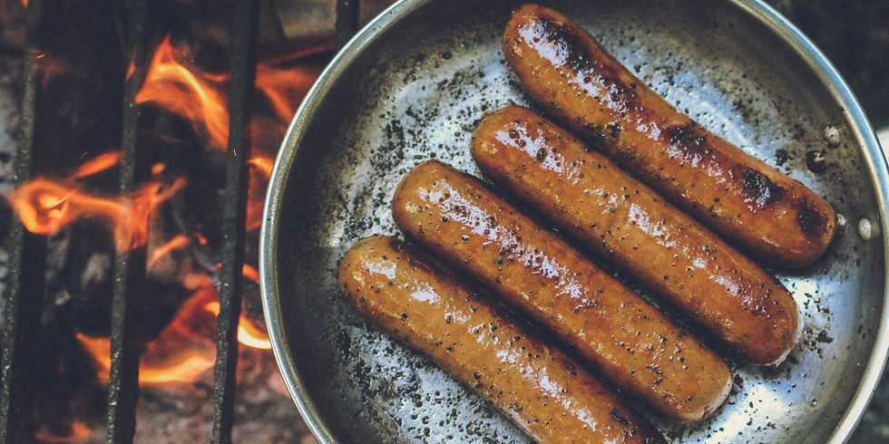Sausages sizzling in a pan over an open flame, emitting a warm glow. The sausages are glossy, with a backdrop of orange and red fire.