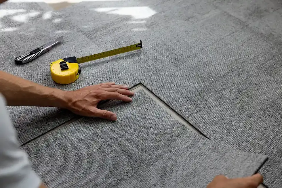 Hands fitting gray carpet tiles, with a yellow tape measure and utility knife nearby on a light surface. Natural lighting creates a calm setting.