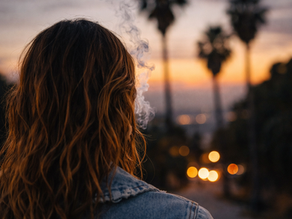 Back view of a young woman with wavy hair in a denim jacket, standing outdoors at dusk with palm trees and soft city lights, as smoke drifts upward into the evening sky.