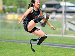 Athlete with a prosthetic leg mid-air during a long jump on a sunny track field. Wearing a black uniform, showing determination.