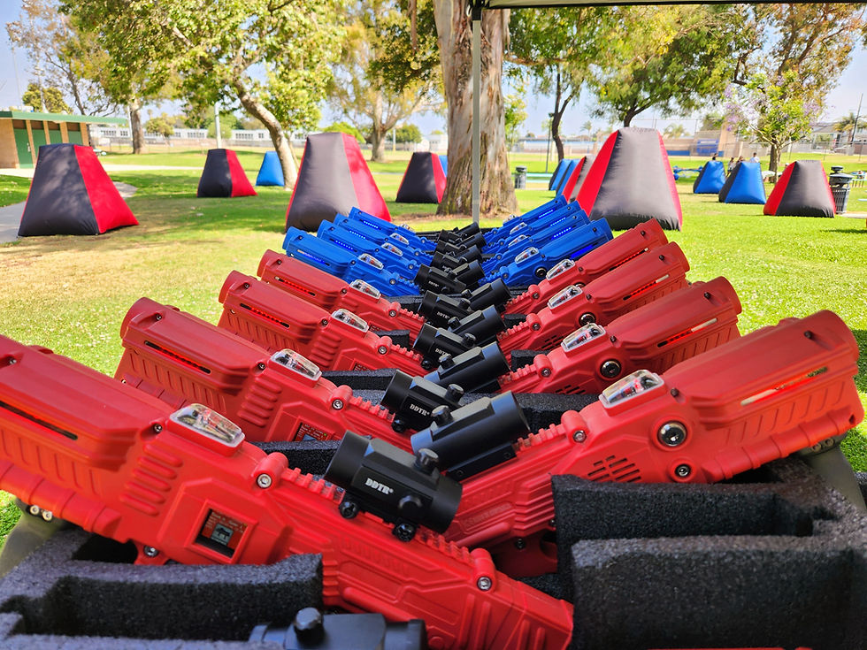 Alt text: Close-up of neatly arranged red and blue laser tag guns on foam racks at an outdoor laser tag arena. In the background, inflatable bunkers in red, blue, and black are scattered across a grassy field with trees and park facilities visible, indicating a recreational activity setup on a sunny day.