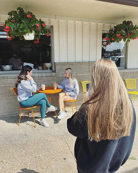 Two people converse at an outdoor cafe table as a third person approaches
