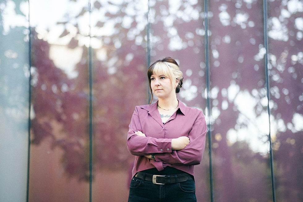 Schauspielportrait in Leipzig mit Marika Braun: Schauspielerin steht vor einer verglästerten Wand, in welcher sich Bäume spiegeln. Die Frau trägt eine altrosafarbene Bluse und hat die Arme vorm Körper verschränkt.