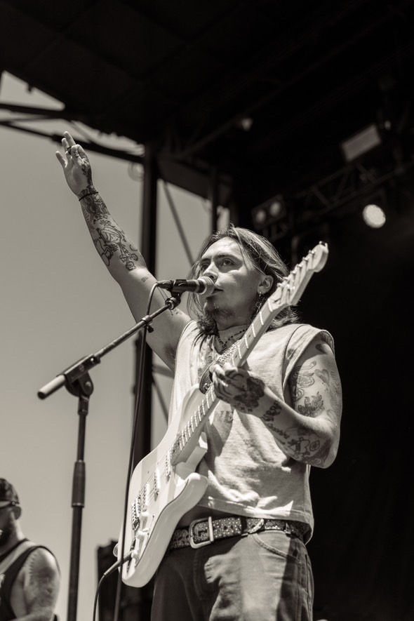 Tattooed musician with a guitar raises hand on stage, singing into a mic. Black and white image, energetic concert vibe.