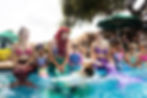 Children delightfully swim alongside the mermaids, with one child playfully splashing about in the water. Ariel, the mermaid princess, looks on with a warm smile, enjoying the magical interaction with the young guests during the mermaid-themed birthday party
