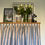 Thumbnail: Putty and blue striped kitchen curtain valance with a pleated design. The valance is made of a fabric with vertical stripes