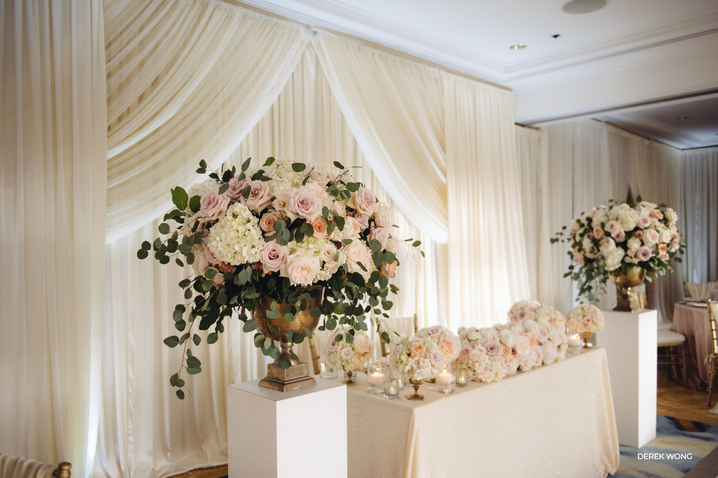 Elegant wedding sweetheart table adorned with soft blush and ivory floral arrangements, cascading greenery, glowing candlelight, and romantic ivory draping that creates a timeless ballroom backdrop.