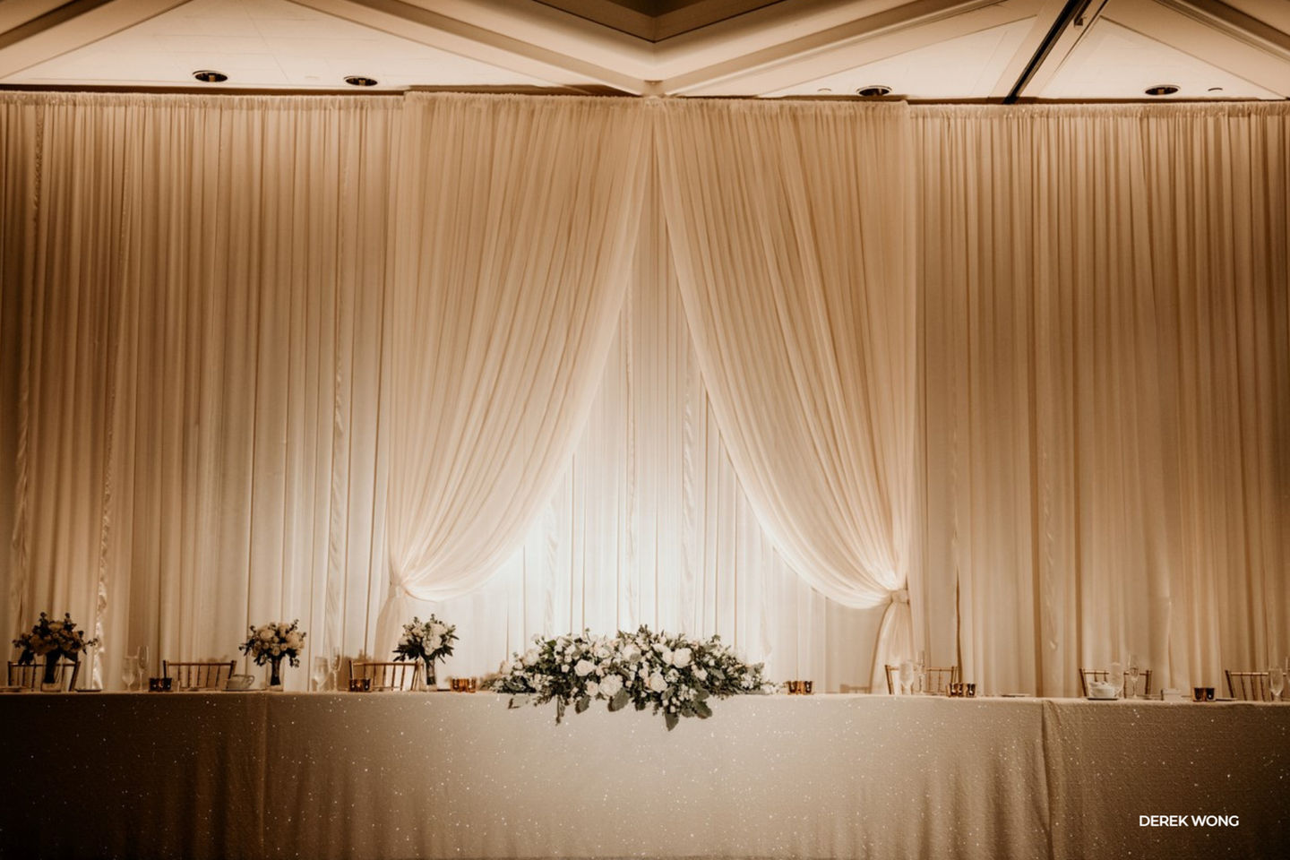 Classic sweetheart table set against a floor-to-ceiling ivory draped backdrop with soft uplighting, featuring a sparkling linen, low white floral arrangement, and gold accents in an elegant ballroom setting.