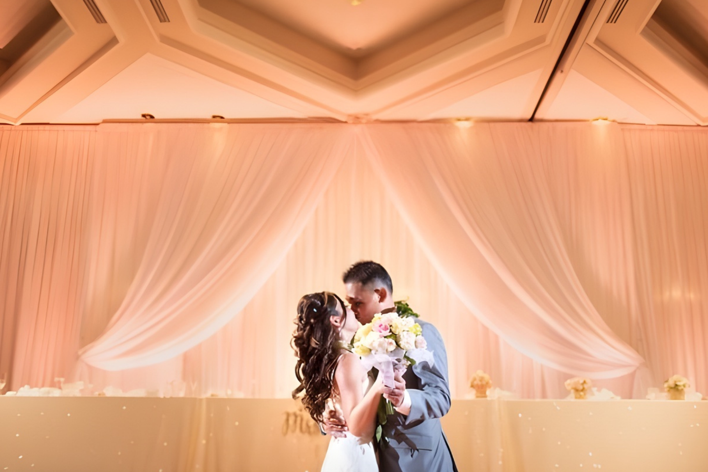Bride and groom share a romantic first dance beneath soft ivory draping and warm uplighting, with a glowing sweetheart table and elegant ballroom details creating a timeless wedding atmosphere.
